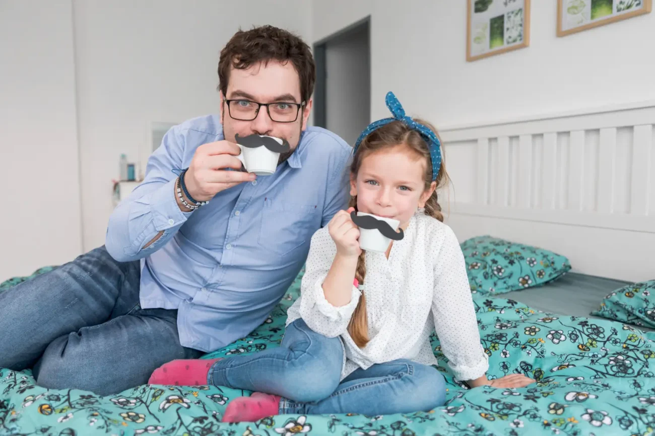 A father and daughter playfully posing with hands near their faces while lying on a bed.
