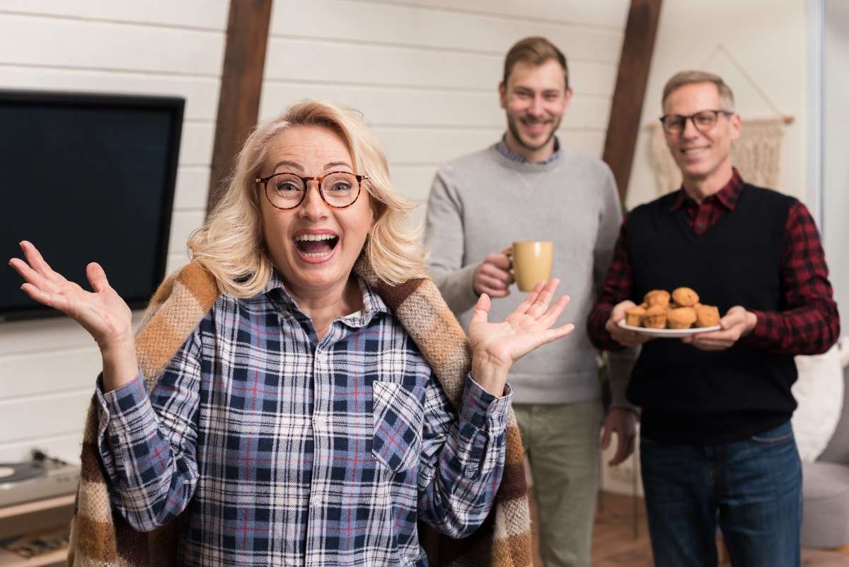 Happy elderly woman smiling and gesturing with excitement while two men stand behind her, one holding a mug and the other a plate of muffins in a cozy living room.