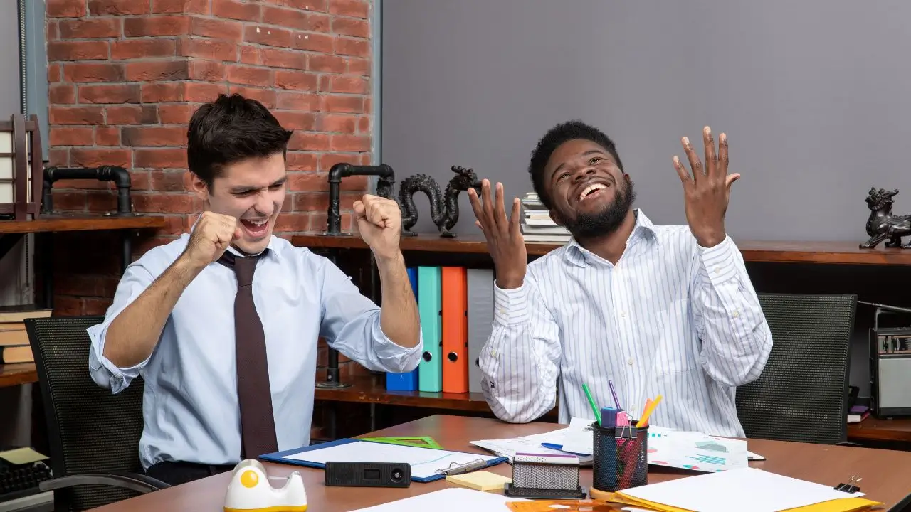 Two office colleagues celebrating and laughing at their desk, showing excitement and joy inspired by funny jokes that’ll make you laugh out loud.