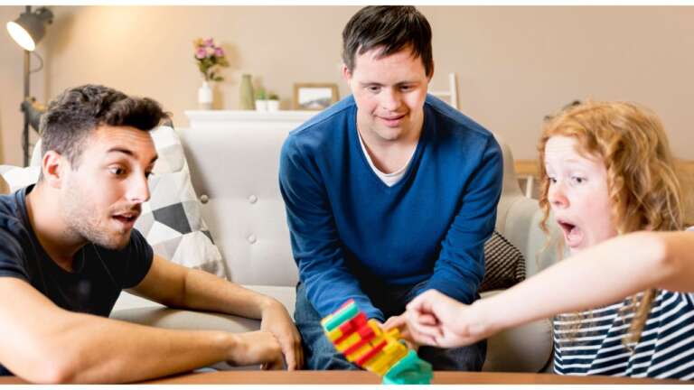 Friends laughing and playing a colorful stacking game together at home, having fun and sharing smiles while solving funny riddles for kids and adults alike.