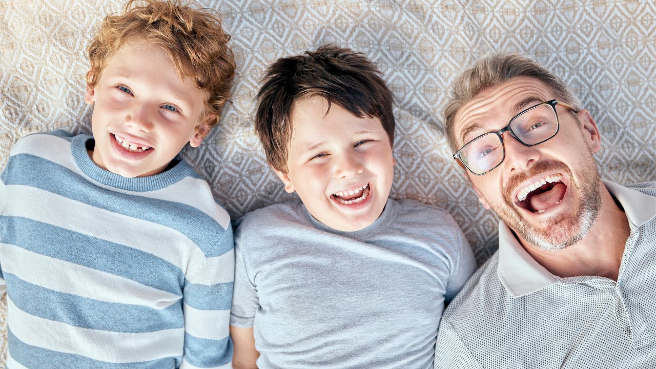 A dad and his two sons smiling widely while lying on the floor, sharing a lighthearted moment. Hilarious Dad Jokes
