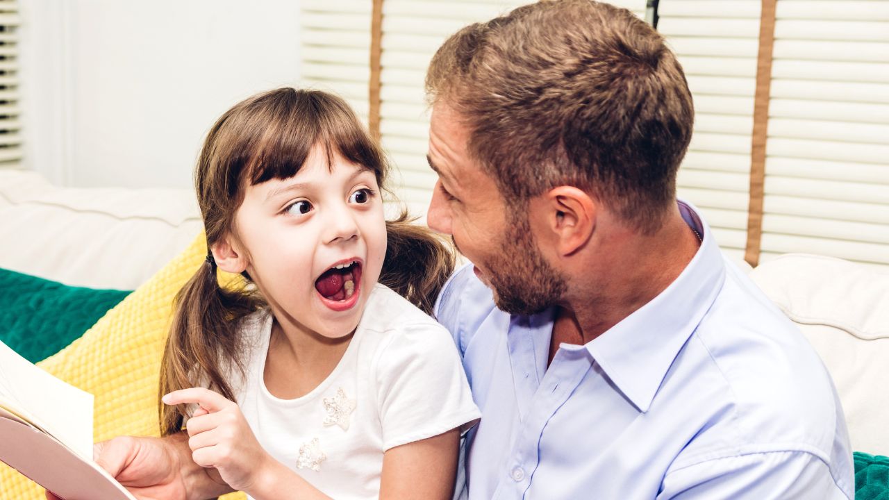 A dad and his daughter laughing together while sitting on a couch, bonding over a joke. Classic Dad Jokes That Never Get Old