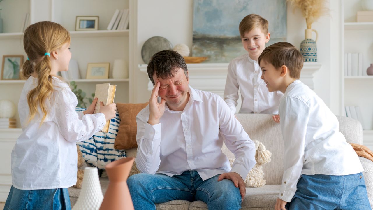 A dad with a slight headache surrounded by his four kids who are playfully teasing him. Family-Friendly Dad Jokes That Bring Everyone Together