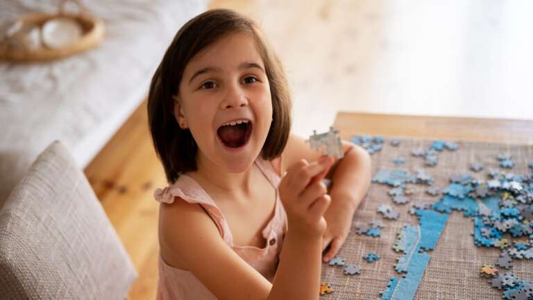 Young girl holding a puzzle piece excitedly while working on a jigsaw puzzle. Fun and Easy Riddles Every Kid Will Love.