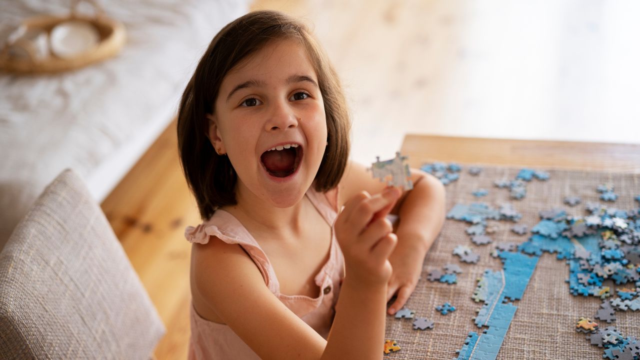 Young girl holding a puzzle piece excitedly while working on a jigsaw puzzle. Fun and Easy Riddles Every Kid Will Love.