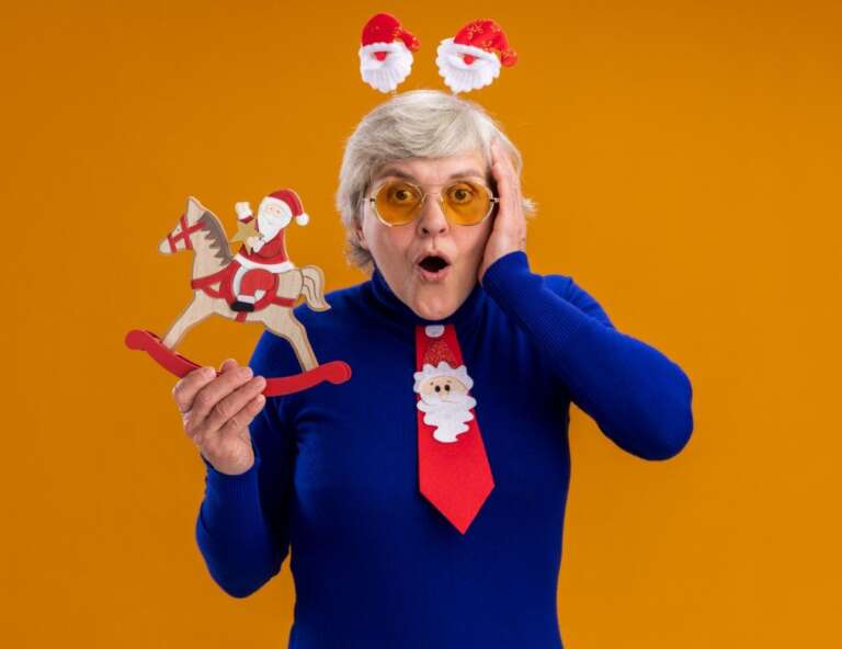 Surprised elderly woman wearing Santa headband and Christmas tie, holding a festive decoration. Brain-Tickling Funny Riddles with Answers – Grandma’s Shocked Reaction to the PunchlineBrain-Tickling Funny Riddles with Answers