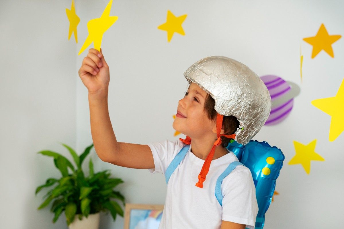 A smiling child wearing a homemade rocket costume and silver helmet while touching paper stars on the wall.