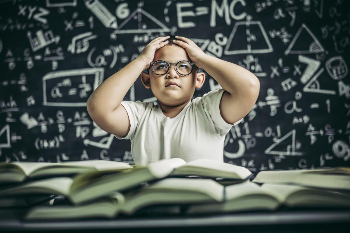A young boy wearing glasses holding his head in confusion while studying with many open books and math formulas on the chalkboard behind him.