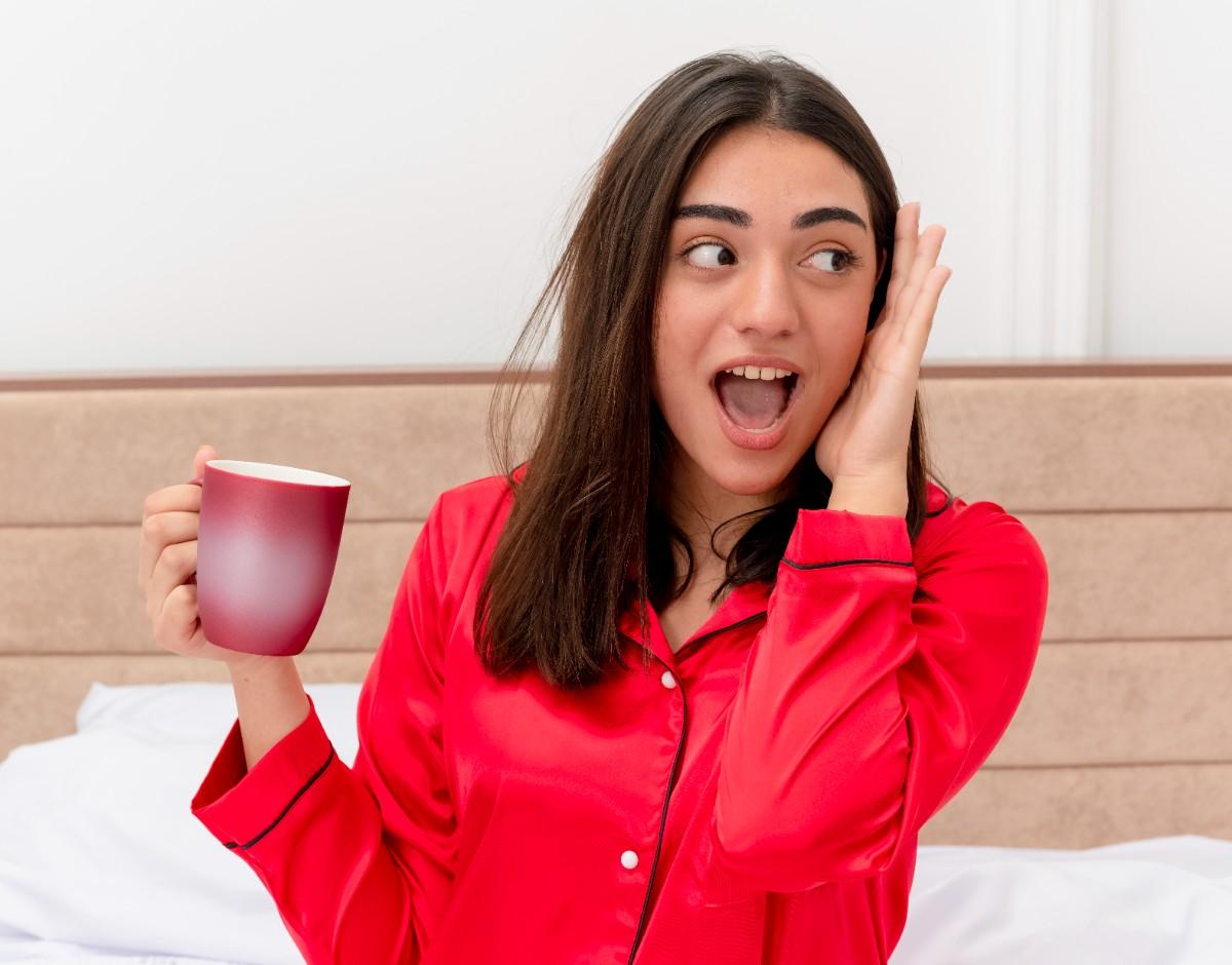 A surprised young woman in red pajamas holding a mug and reacting excitedly.