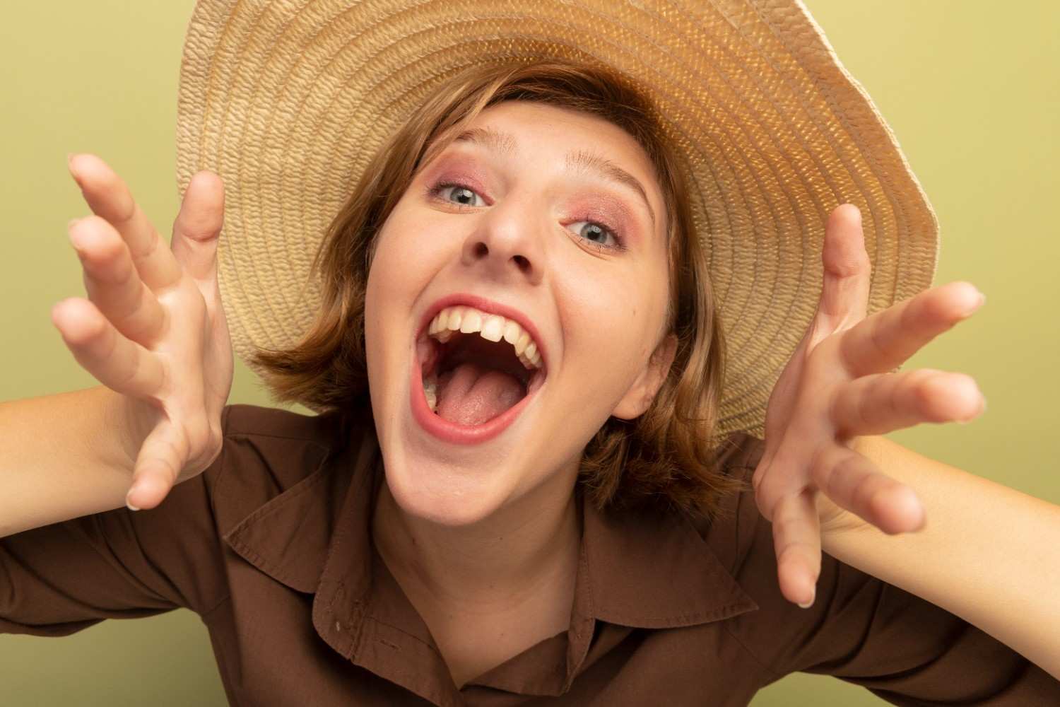 A cheerful woman wearing a big straw hat laughing loudly with hands raised playfully.