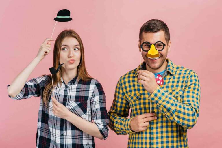 Two people posing with funny props, wearing playful disguises and smiling on a pink background.