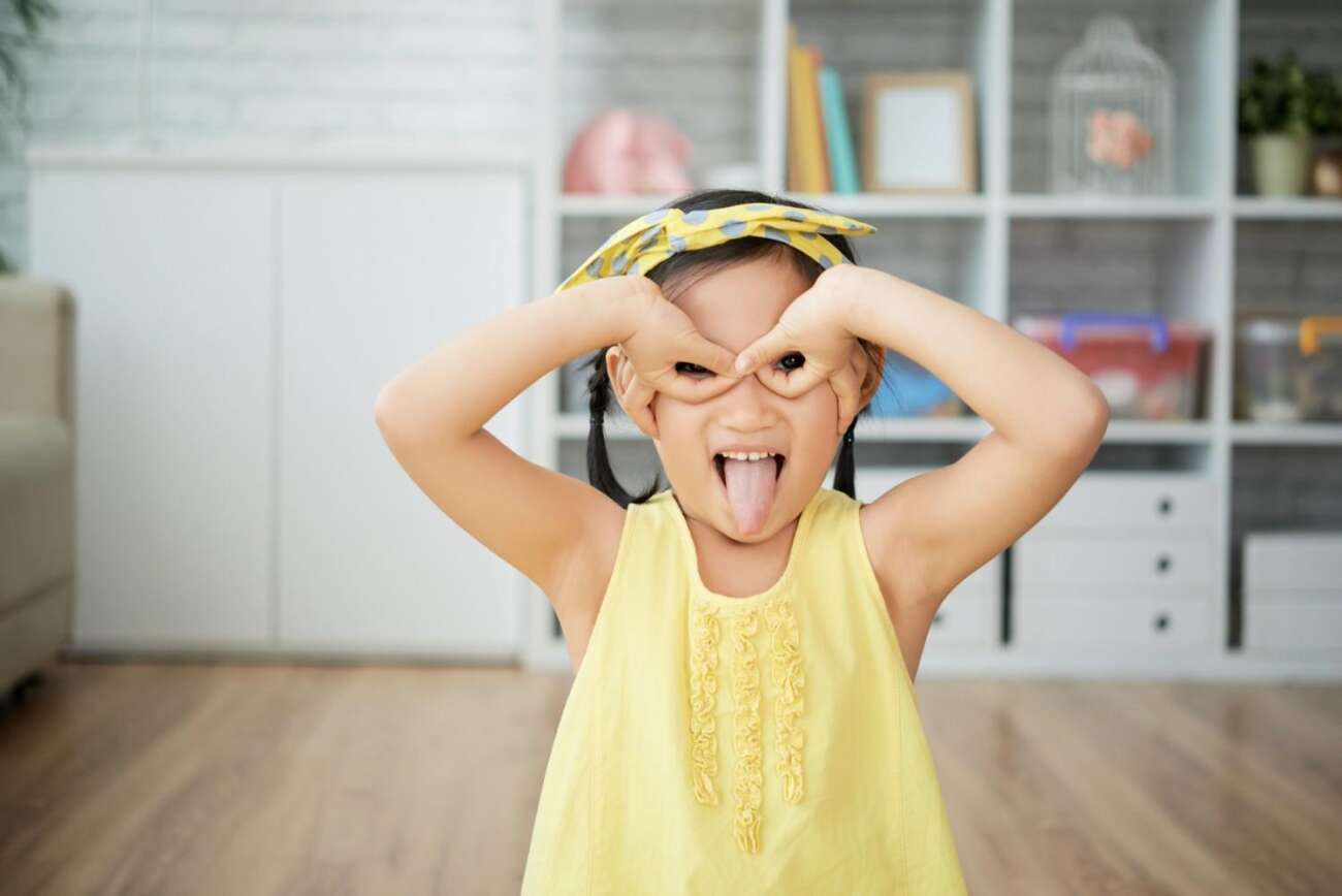 A happy little girl making a silly face with her tongue out and hands shaped like glasses over her eyes.