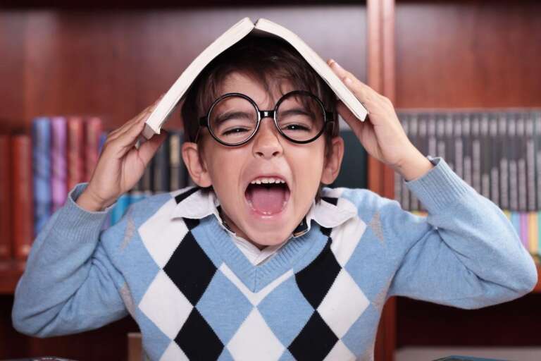 A young boy with glasses screams in frustration while holding a book above his head, set against a bookshelf. Kid-Friendly Funny Jokes You Can’t Read Without Laughing – The Hilarious Struggle of Reading.