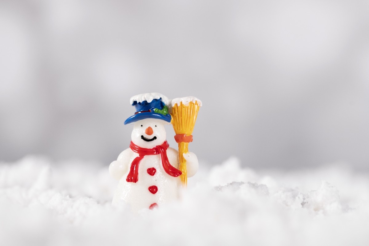 A small, cute snowman with a blue hat and red scarf standing amidst snowy white decoration. Best Christmas Jokes That’ll Get Everyone Laughing This Holiday: Cheerful Snowman with Christmas Vibes.