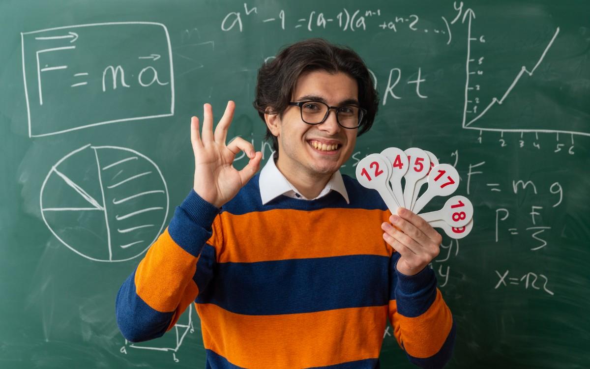 A young smiling teacher in glasses holding number cards in front of a chalkboard filled with math and science equations. Funny School Jokes That’ll Make You Miss Class (Almost!)